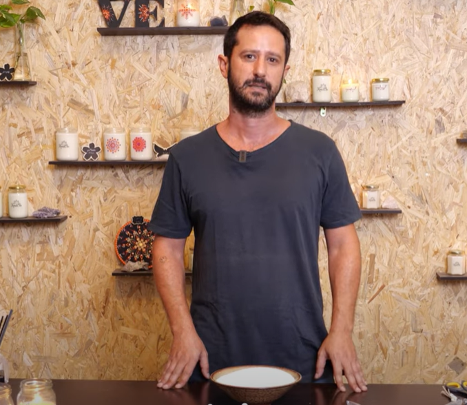אישה עומדת מול שולחן עם קערה, ברקע מדפים עם נרות ריחניים ופריטי דקורציה